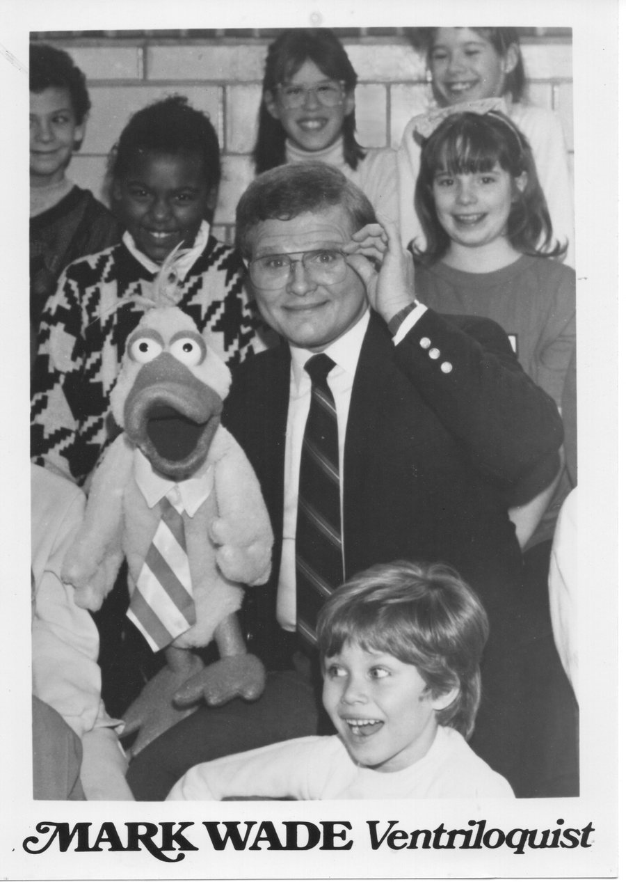 Black and white publicity photo of Mark Wade surrounded by schoolchildren, holding a bird figure