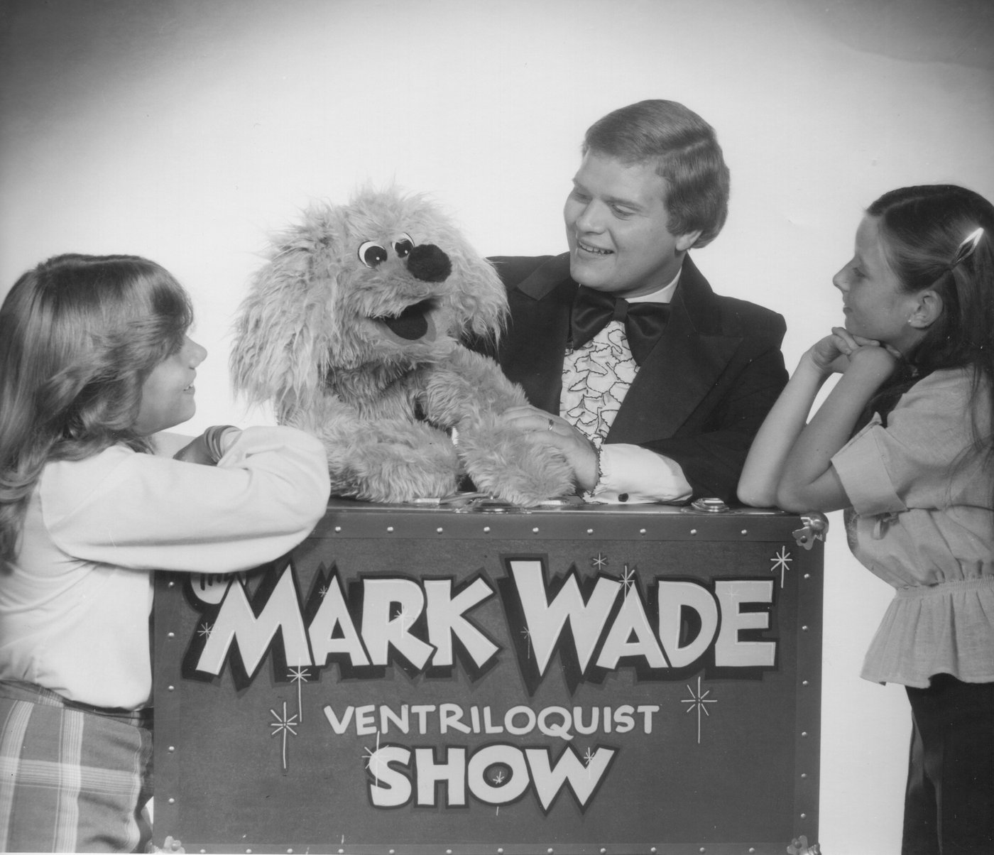 A young Mark Wade in a tuxedo with a shaggy dog puppet and two children, leaning on a trunk painted “The Mark Wade Ventriloquist Show”